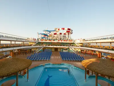 Weitwinkelansicht eines leeren Pooldecks auf der Carnival Horizon. Blaue Liegestühle und zwei große Wasserrutschen, eine rot-weiß, eine blau-weiß, sind zu sehen. Im Vordergrund ein Pool mit Delfinschwanz-Muster. Bars und weitere Decks umgeben den Bereich.