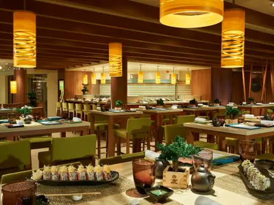 Innenansicht eines eleganten, japanischen Spezialitätenrestaurants mit langen Holztischen, grünen Stühlen, warmen Hängelampen und einer Sushi-Bar im Hintergrund. Im Vordergrund liegt Sushi auf einem Teller.