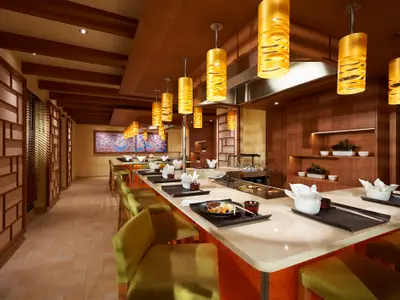 Ein modern eingerichtetes Teppanyaki-Restaurant an Bord der Carnival Horizon. Eine lange Theke mit hellen Arbeitsplatten erstreckt sich durch den Raum, bestückt mit Tellern und Essbesteck. Grüne Barhocker stehen davor. Im Hintergrund ist eine japanische Kirschblütenmalerei zu sehen. Die Decke ist mit Holzbalken und vielen goldfarbenen Hängelampen verziert, die eine warme Atmosphäre schaffen.