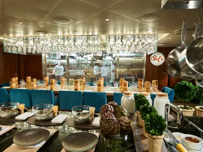 Elegantes Spezialitätenrestaurant mit langem Tisch, türkisen Stühlen und Blick in die offene Küche mit Köchen. Über dem Tisch eine Lichtinstallation mit Gläsern. Rechts ein Schild 'The Chef's Table'.