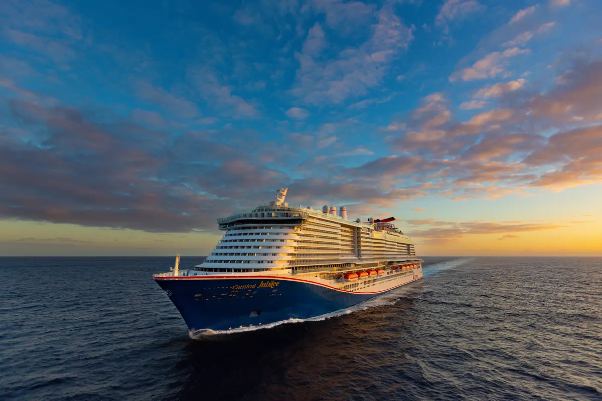 Die moderne Carnival Jubilee fährt bei Sonnenuntergang auf dem offenen Meer. Der blaue Rumpf und das weiße Deck sind beleuchtet. Der Himmel zeigt rosa-orange Wolken.