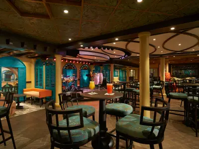 Eine lebhafte Bar-Lounge auf der Carnival Jubilee mit einer Bühne, beleuchteten Musikinstrumenten und zwei Cocktails auf einem runden Tisch in der Mitte des Raumes. Die Einrichtung ist farbenfroh, mit türkisfarbenen Fensterläden und goldgelben Wänden. Die Decke hat kunstvolle Ornamente und Lichter.