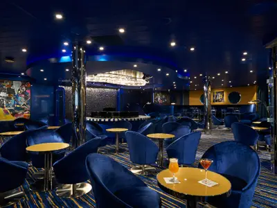 Bar-Lounge auf der Carnival Jubilee in tiefem Blau, mit Samtstühlen und Goldtischen. Ein Musiker-Gemälde links und ein Kronleuchter prägen den Raum. Zwei Cocktails im Vordergrund.