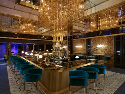 Eine luxuriöse Bar-Lounge auf der Carnival Jubilee mit einer U-förmigen Theke aus dunklem Marmor und goldenen Akzenten. Blaue Samt-Barhocker stehen bereit, und eine opulente goldene Lichtinstallation hängt von der Decke. Mehrere Cocktails sind auf der Theke zu sehen.