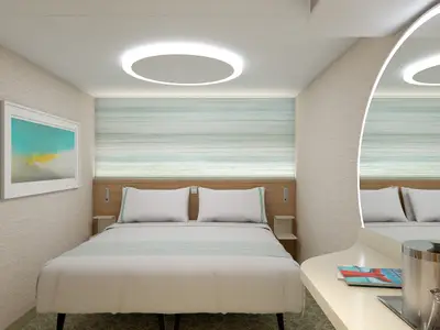 Eine helle, modern gestaltete Kreuzfahrtkabine mit Doppelbett, weißen Kissen und blauer Tagesdecke. Eine abstrakte Wanddekoration und indirekte Deckenbeleuchtung prägen das Ambiente. Rechts ist ein großer Spiegel sichtbar.