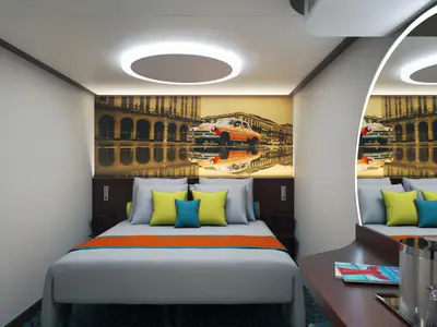 Moderne Kabine der Carnival Jubilee mit Doppelbett, hölzernen Nachttischen. Wandkunst mit orangem Oldtimer und Reflexionen über dem Bett. Bunte Kissen und Deckenbeleuchtung.
