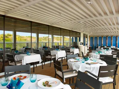 Ein elegantes Restaurant auf der Carnival Jubilee mit vielen Tischen, weiß eingedeckt, blauen Gläsern und Stühlen. Große Fensterfronten bieten einen Ausblick auf grüne Hügel. Die Decke ist mit weißen Lamellen verziert.