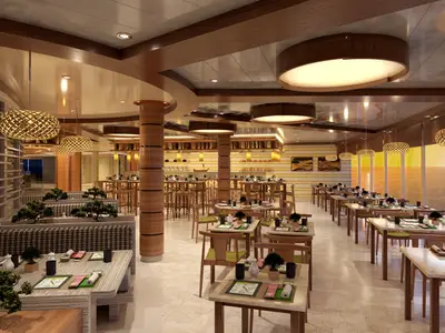 Innenansicht eines stilvollen, modernen asiatischen Spezialitätenrestaurants auf der Carnival Jubilee. Zahlreiche gedeckte Tische mit Blick auf das Meer bei Sonnenuntergang. Helle Holz- und Naturmaterialien dominieren das Design, ergänzt durch dekorative Bonsaibäume und auffällige Hängelampen.