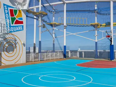 Ein leeres Sportdeck auf der Carnival Jubilee an einem sonnigen Tag, mit einem blauen Basketballfeld, einem roten Freiwurfbereich und einem dahinter liegenden Hochseilgarten.