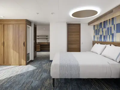 Helle, moderne Kreuzfahrtkabine mit Doppelbett, weißer Bettwäsche, blauen Akzenten, Holzwänden und einem separaten Ankleidebereich.