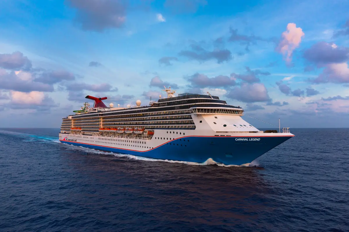 Luftaufnahme der Carnival Legend, die bei leicht bewölktem Himmel und Sonnenuntergang auf blauem Meer fährt. Das Schiff ist weiß, blau und rot.