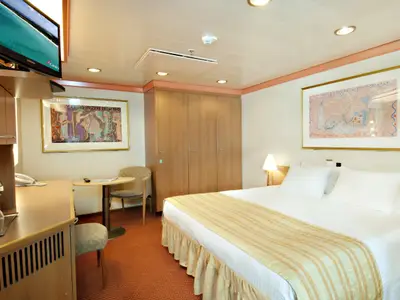 Geräumige und helle Außenkabine auf der Carnival Legend mit einem gemütlichen Doppelbett, einem Schreibtischbereich und einem großen Kleiderschrank aus Holz. Zwei gerahmte Kunstwerke schmücken die Wände, und ein Fernseher ist an der Wand montiert.
