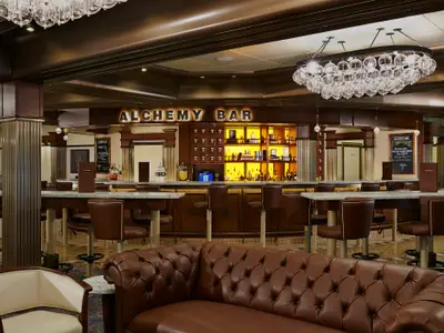Eine stilvolle Bar auf der Carnival Legend mit einem langen Tresen, braunen Barhockern und einem leuchtenden 'ALCHEMY BAR'-Schriftzug. Eine große Kristallleuchte hängt von der Decke, im Vordergrund ein braunes Chesterfield-Sofa.