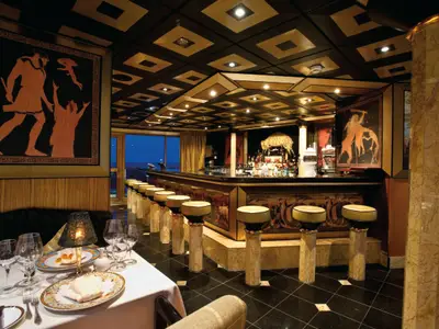 Eine elegante Schiffsbar bei Abendstimmung, mit dunklen Holz- und Marmorelementen. Gedeckte Tische im Vordergrund, eine lange Bar mit Säulen Hockern und einer gut bestückten Rückwand, verziert mit antiken Kunstwerken, führen zu großen Fenstern mit Blick auf das dunkle Meer.