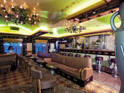 Blick in eine stilvolle Bar und Lounge auf der Carnival Legend. Dunkle Ledersofas und Stühle, eine lange Bar mit Flaschen, und ein Barkeeper. Große Fenster bieten einen weiten Blick auf das offene Meer. Die Decke ist hellgrün mit goldenen Ornamenten und auffälligen Kronleuchtern. Ein großer, urnenartiger Deko-Gegenstand ist rechts im Vordergrund.