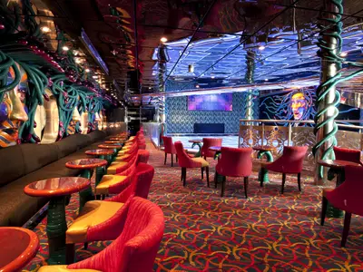 Extravagante Bar-Lounge auf der Carnival Legend. Auffällige Masken und Schlangen-Elemente. Rote Sessel, gemusterter Teppich, blaue Deckenbeleuchtung.