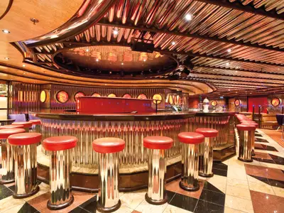Eine Innenansicht einer modernen Bar-Lounge auf der Carnival Legend. Eine lange, geschwungene Bar mit glänzenden, vertikal gerippten Oberflächen erstreckt sich durch den Raum. Zahlreiche rote Barhocker mit silbernen Säulen stehen davor. Im Hintergrund sind runde, leuchtende Fenster und bequeme Sitzgelegenheiten zu sehen. Die Decke ist mit Holz und Metallakzenten verziert.