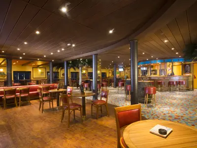 Innenansicht einer geräumigen Bar-Lounge auf der Carnival Legend mit einem langen Tresen, roten Barhockern und Sitzbänken. Das Interieur ist warm beleuchtet, hat Holzdecken und einen bunten Teppichboden. Palmen und Bilder an den Wänden schmücken den Raum.
