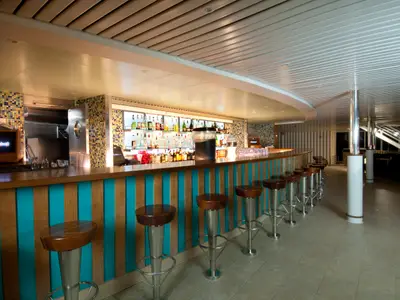 Eine lange, bogenförmige Bar auf der Carnival Legend. Der Tresen hat Holzoberfläche und die Front ist mit abwechselnd türkisen und holzfarbenen vertikalen Streifen verziert. Eine Reihe von braunen Barhockern mit silbernen Füßen steht bereit. Im Hintergrund ist eine große, beleuchtete Spirituosenwand zu sehen, umgeben von farbenfrohen Mosaikfliesen. Ein Bildschirm mit dem 'Carnival'-Logo ist links platziert. Die Decke ist gerippt und hell.