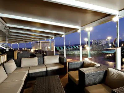 Abend auf dem Oberdeck der Carnival Legend mit bequemen Loungemöbeln unter einem Vordach. Im Hintergrund ein beleuchteter Pool und Liegestühle bei blauer Dämmerung.