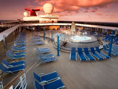 Das Sonnendeck der Carnival Legend bei Sonnenuntergang. Blaue Liegestühle, Pools und ein Whirlpool auf dem Holzdeck. Im Hintergrund das Meer und Teile des Schiffes.