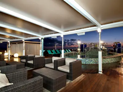 Ein überdachter Poolbereich der Carnival Legend bei Sonnenuntergang. Dunkle Rattansessel stehen um einen mosaikverzierten Pool mit Liegestühlen und warmem Licht.