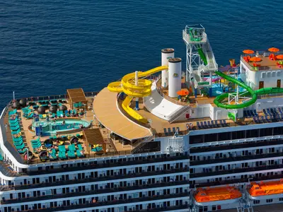 Luftaufnahme des hinteren Sonnendecks der Carnival Legend mit Passagieren, die in einem Pool schwimmen und auf Liegen entspannen. Mehrere farbige Wasserrutschen und Schornsteine sind ebenfalls sichtbar. Das Schiff fährt auf blauem Meer.