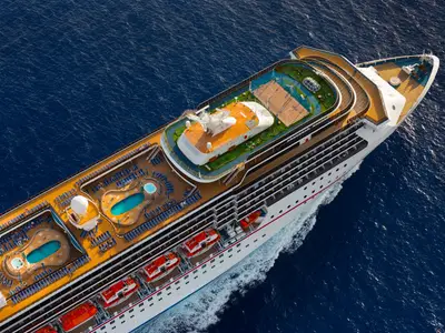 Luftaufnahme der Carnival Legend auf dem Meer. Die oberen Decks zeigen Pools, Liegestühle, einen Minigolfplatz und einen Sportplatz. Blaues Wasser umgibt das Schiff.