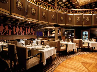 Innenansicht eines eleganten Spezialitätenrestaurants mit vielen gedeckten Tischen und Stühlen. An den Wänden sind antike Motive und goldene Masken zu sehen. Ein Bartresen ist im Hintergrund erkennbar.