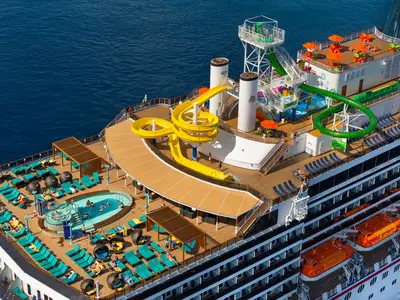 Blick von oben auf das Sonnendeck der Carnival Legend. Gäste entspannen an Pools, während Kinder die gelben und grünen Wasserrutschen nutzen. Das Meer ist tiefblau.