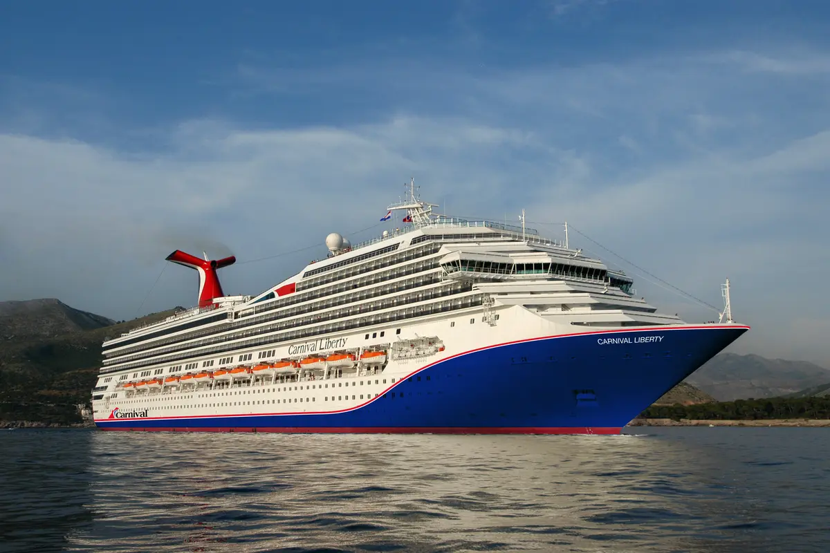 Die große Carnival Liberty mit blauem Rumpf und rotem Schornstein fährt auf dem Meer. Im Hintergrund sind grüne Berge und ein blauer Himmel zu sehen.