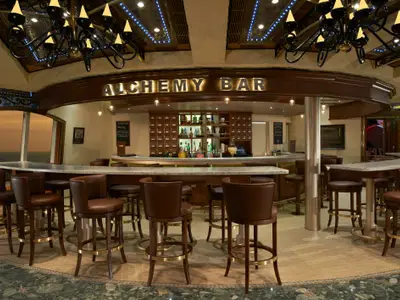Die Alchemy Bar auf der Carnival Liberty. Die halbkreisförmige Bar mit hohen Hockern und braunen Ledersitzen blickt auf das Meer bei Sonnenuntergang. Der Tresen ist mit Flaschen und Gläsern bestückt. Elegantes Interieur mit dunklem Holz und Lichtern.