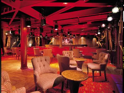 Eine luxuriöse Bar-Lounge mit tiefroter Beleuchtung und Plüschmöbeln. Links und rechts der Bar stehen kunstvolle Holzstatuen. Die Decke ist rot gestrichen und mit vielen Strahlern bestückt. Ein Bartresen ist im Hintergrund zu sehen.