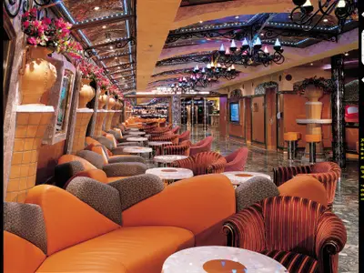 Weitwinkelansicht einer opulenten Bar-Lounge auf der Carnival Liberty. Die Szene ist gefuellt mit Reihen von orangefarbenen Sesseln, runden Tischen, dunklen Holzvertäfelungen und kunstvollen Deckenleuchten mit bunten Lichtern.