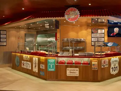 Ein lebhafter Burger-Joint im Diner-Stil auf der Carnival Liberty, mit einer langen Theke, Menütafeln, frischen Burgerpatties und einem Fernseher, der Guy Fieri zeigt.