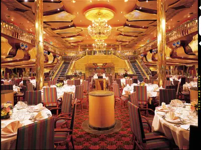 Innenansicht eines opulenten Restaurants auf der Carnival Liberty. Mehrere festlich gedeckte Tische mit bunten Stühlen sind zu sehen, umgeben von glänzenden goldenen Säulen, Deckenornamenten und einer breiten Doppeltreppe. Die Beleuchtung ist warm und einladend.