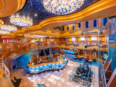 Ein weitläufiges Atrium der Carnival Luminosa mit einer beleuchteten Bar, Marmorböden und geschwungenen Treppen. Deckenleuchten mit blauen Glaselementen hängen über einem bronzefarbenen Wanddesign. Ein Mann entspannt auf einem Sessel, nahe einer modernen Skulptur einer liegenden Frau.