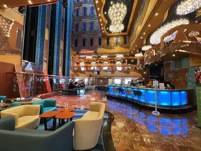 Innenansicht eines mehrstöckigen Atriums an Bord der Carnival Luminosa, aufgenommen in der Dämmerung oder am Abend. Eine langgezogene, blau beleuchtete Bar mit Hockern dominiert den Vordergrund. Davor und daneben befinden sich bequeme Sitzgruppen in hellen Farben. Im Hintergrund erheben sich mehrere Ebenen mit kunstvollen Wänden in Gold- und Brauntönen, geschmückt mit glitzernden Kronleuchtern. Eine dunkle Skulptur liegt auf einem Podest links. Links sind die reflektierenden Wände eines Aufzugs sichtbar.