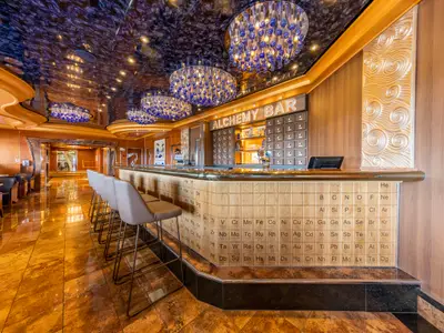 Eine moderne Bar auf der Carnival Luminosa, genannt 'Alchemy Bar', mit einer Theke, die mit Elementen des Periodensystems verziert ist. Blaue Glasleuchten hängen von der glänzenden Decke.