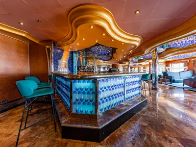 Moderne Bar-Lounge auf der Carnival Luminosa. Blaue Leuchttheke, türkise Barhocker. Goldene, geschwungene Decke mit blauen Kronleuchtern und Lichtern. Blaue Loungesessel, glaenzender Boden.