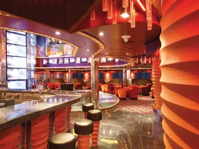 Blick in eine moderne, farbenfrohe Bar und Lounge auf der Carnival Luminosa. Links eine lange Bar mit dunkler Granitplatte und Barhockern. Rechts bequeme rote und orange Sessel sowie eine große, gewellte orangefarbene Säule. Der Raum ist hell beleuchtet und hat einen glänzenden dunklen Boden.