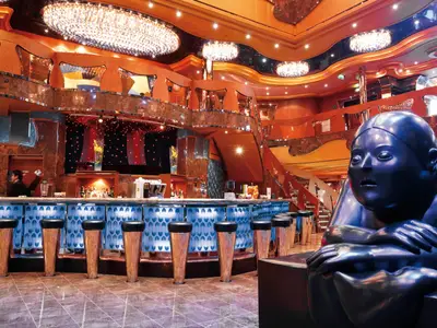 Innenansicht einer opulenten Bar der Carnival Luminosa mit blauer Leuchttheke, vielen Barhockern, einer Bartenderin und einer großen, dunklen Skulptur im Vordergrund. Mehrere Ebenen und Kronleuchter im Hintergrund.
