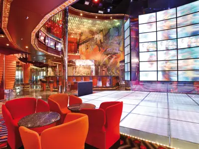 Ansicht einer lebhaften Lounge und Clubbereich auf der Carnival Luminosa. Die Szene zeigt rote Sessel, eine Bar, einen beleuchteten Tanzboden und eine Videowand mit abstrakten Bildern.