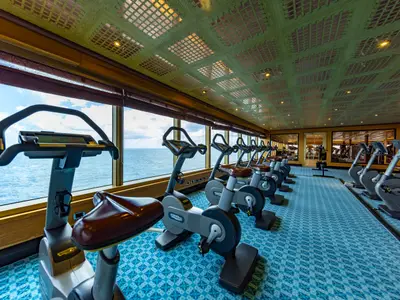 Ein breiter Blick in ein Fitnessstudio auf der Carnival Luminosa. Mehrere stationäre Fahrradergometer stehen vor großen Fenstern mit Blick auf das Meer. Der Raum hat einen gemusterten blauen Teppich und eine Gitterdecke.