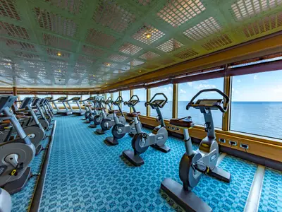 Ein helles Fitnessstudio auf der Carnival Luminosa mit einer Reihe grauer Technogym-Ergometer und Laufbänder. Große Fensterfront bietet einen weiten Blick auf das blaue Meer und den Himmel. Der Boden ist mit blauem Teppich ausgelegt.