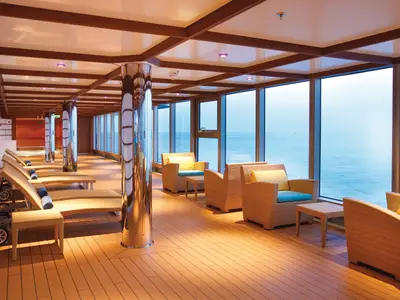 Blick in eine moderne Schiffs-Lounge mit bodentiefen Fenstern und weitem Meerblick. Im Vordergrund stehen Liegestühle und Korbsessel auf einem Holzdeck. Die Decke hat eine Holzgitterstruktur.