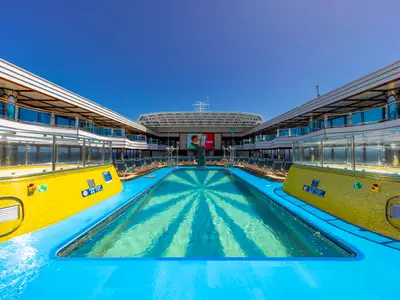 Weitwinkelaufnahme des menschenleeren Pooldecks der Carnival Luminosa an einem sonnigen Tag. Ein großer Pool mit Muster im Wasser, umgeben von gelben Fliesen, erstreckt sich zur Leinwand am Heck. Sonnenliegen stehen bereit unter mehrstoeckigen Decks.