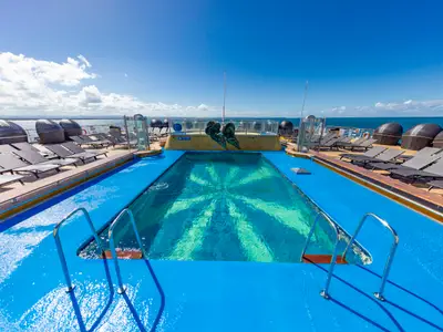 Sonniges, menschenleeres Sonnendeck der Carnival Luminosa. Im Vordergrund ein blauer Pool mit Muster, umgeben von Liegestühlen und Cabanas. Dahinter Skulptur, offenes Meer und blauer Himmel.