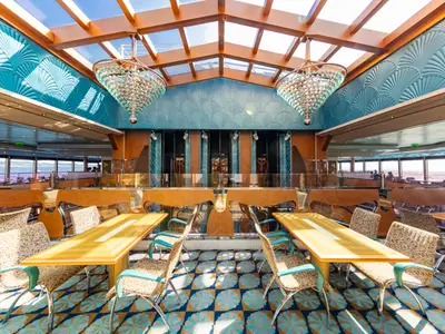 Ein elegantes Restaurant auf der Carnival Luminosa mit hohen Decken und großen Fenstern. Türkisfarbene, fächerförmige Wandpaneele und glitzernde Kronleuchter prägen das art-deco-inspirierte Design. Zahlreiche Tische und Stühle laden zum Verweilen ein, während man den Meerblick genießen kann.