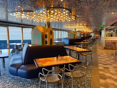 Blick in ein modernes Schiffsrestaurant mit großen Fenstern zum Meer. Blaue Sitzbänke, Tische, Stühle, verspielte Haus-Dekoration und eine leuchtende Deckeninstallation.